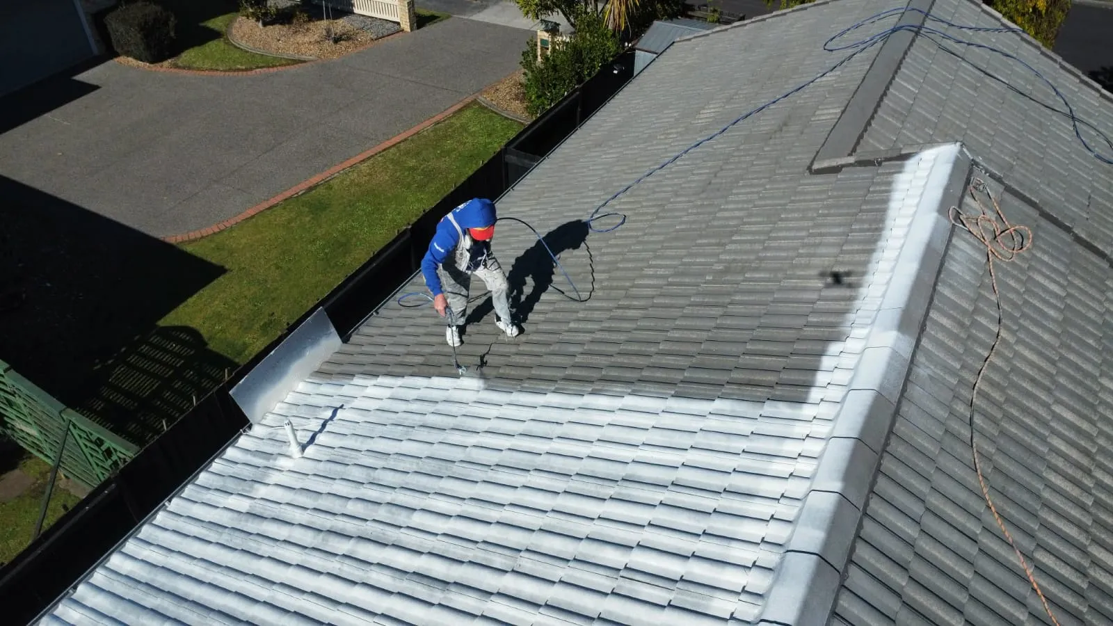 A roofer in a harness spraying sealer across a tile roof on a clear Waikato day